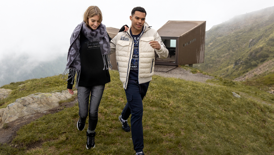 Ein Paar steht auf einem gepflasterten Weg. Die Frau trägt ein pinkes Oberteil und eine weiße Hose, während der Mann ein blaues Hemd über einem weißen T-Shirt und hellen Jeans trägt.