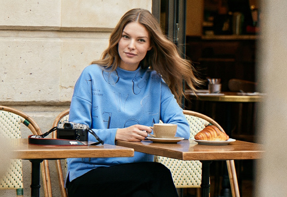 Model sitzt im Cafe und trägt blauen SOCCX Pullover