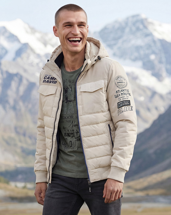 Model steht in einer Berglandschaft und trägt eine weiße Jacke